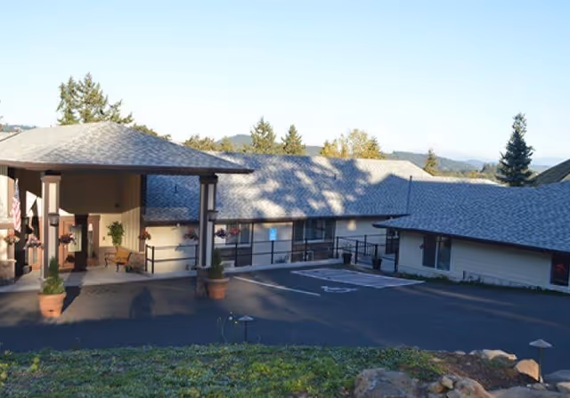 Front entrance and driveway of a single-story senior living building with a covered porte-cochère, potted plants, and surrounding landscaping.