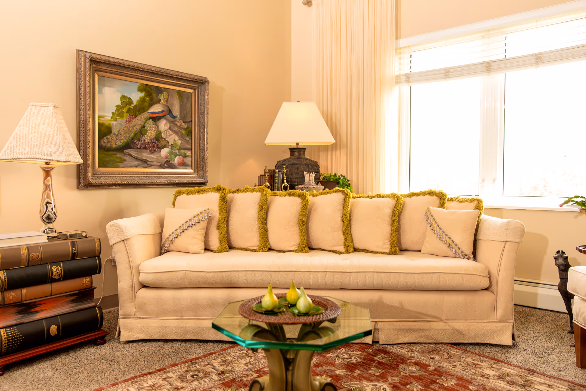 A cozy living room with a beige sofa adorned with multiple cushions with green fringes, a glass coffee table with a decorative plate holding pears, a side table with a lamp shaped like stacked books, a framed painting of a peacock on the wall, and a large window with blinds letting in natural light.