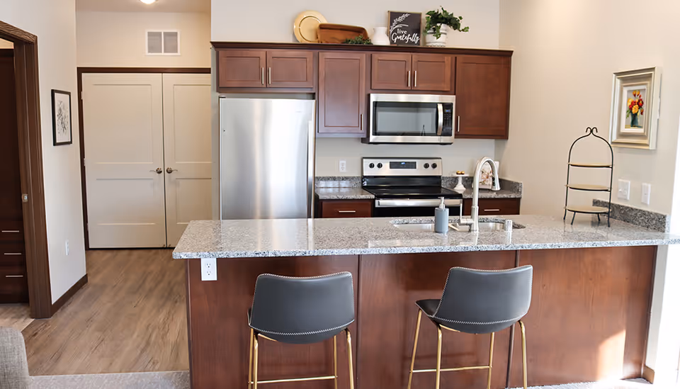 Modern kitchen with granite countertop island, two gray bar stools, stainless steel refrigerator, stove, and microwave, wooden cabinets, and decorative items on top of the cabinets and walls.