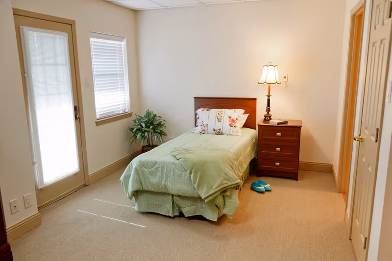 A small, neatly arranged bedroom with a single bed covered in a light green blanket and floral pillows. Next to the bed is a wooden nightstand with a lamp and a book. There is a pair of blue slippers on the beige carpet floor. A potted plant is placed near a window with blinds, and a door with a window is on the left side of the room.