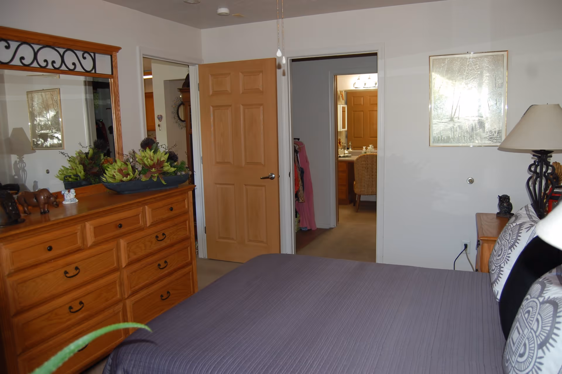 A bedroom with a wooden dresser topped with decorative plants and figurines, a large mirror above the dresser, a bed with a gray quilt and patterned pillows, a bedside table with a lamp and owl figurine, and two open doors leading to another room and a closet.