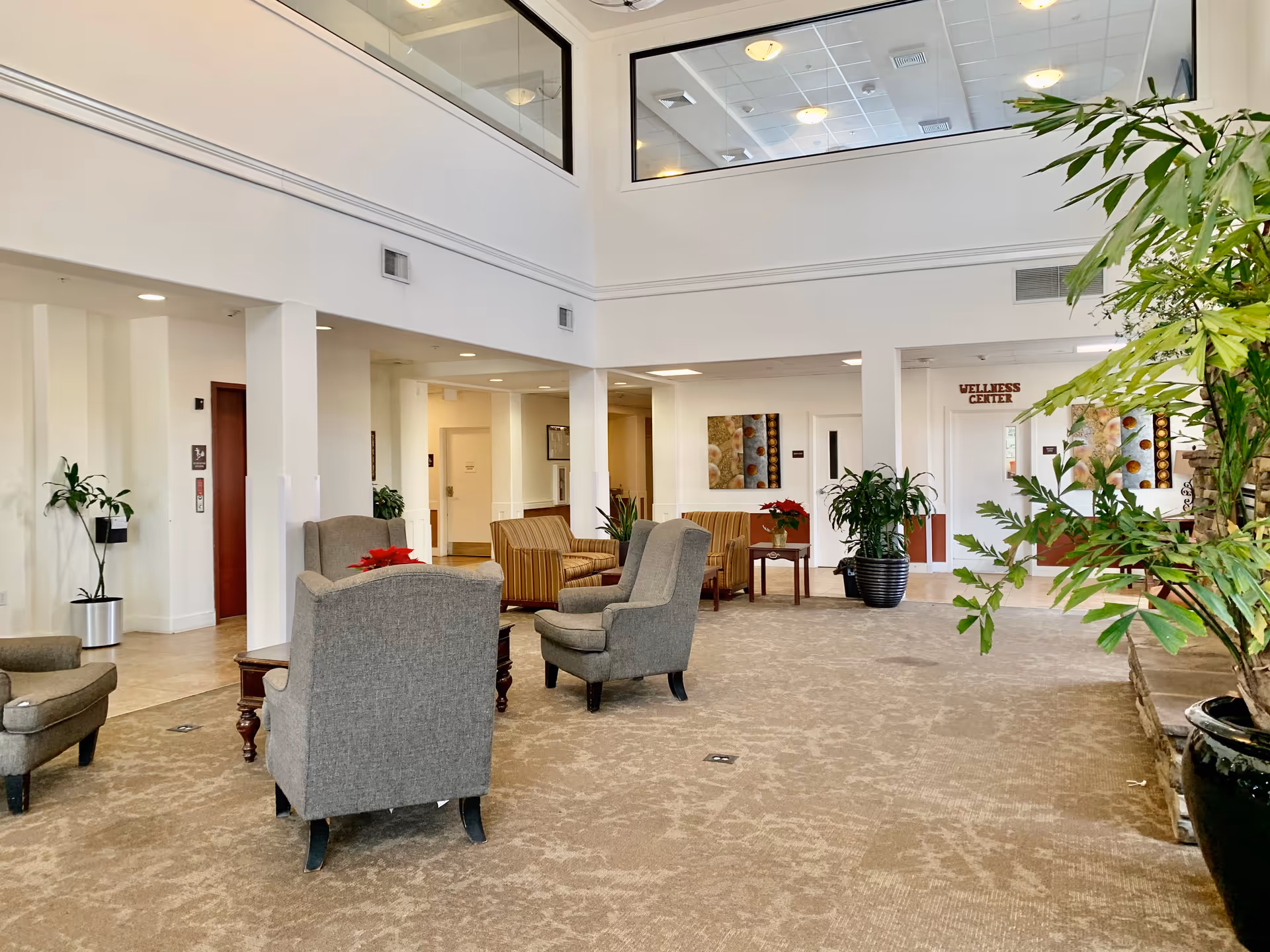A spacious and well-lit common area in an assisted living facility with comfortable armchairs and sofas arranged around small tables. The room features large windows near the ceiling, potted plants, and a sign indicating the Wellness Center in the background.