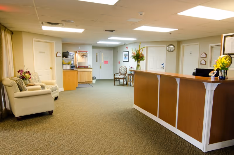 Reception area of Westward Heights Care Center featuring a wooden front desk with flowers on top, beige armchairs with cushions on the left, a popcorn machine in the back, and several doors along the walls under fluorescent lighting.