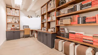 Bright interior study with long built-in wooden shelves filled with books and baskets, a desk and chair beneath an exposed-beam ceiling.