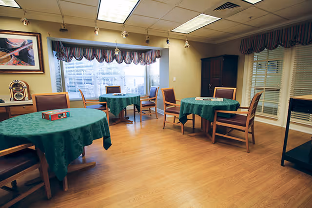 A cozy room with three round tables covered with green tablecloths, each surrounded by wooden chairs with cushions. The room has wooden flooring, a large window with striped valance curtains, and framed artwork on the wall. There is a small cabinet and a cart in the corner.