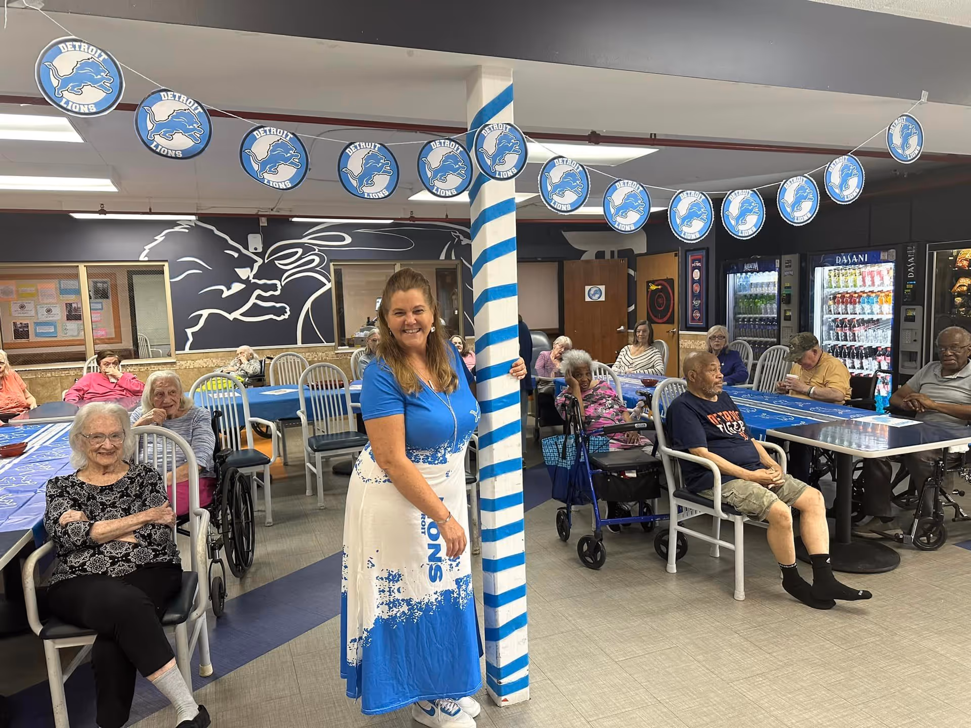 A group of senior residents seated around tables in a decorated activity/dining room with a staff member standing by a striped pole.