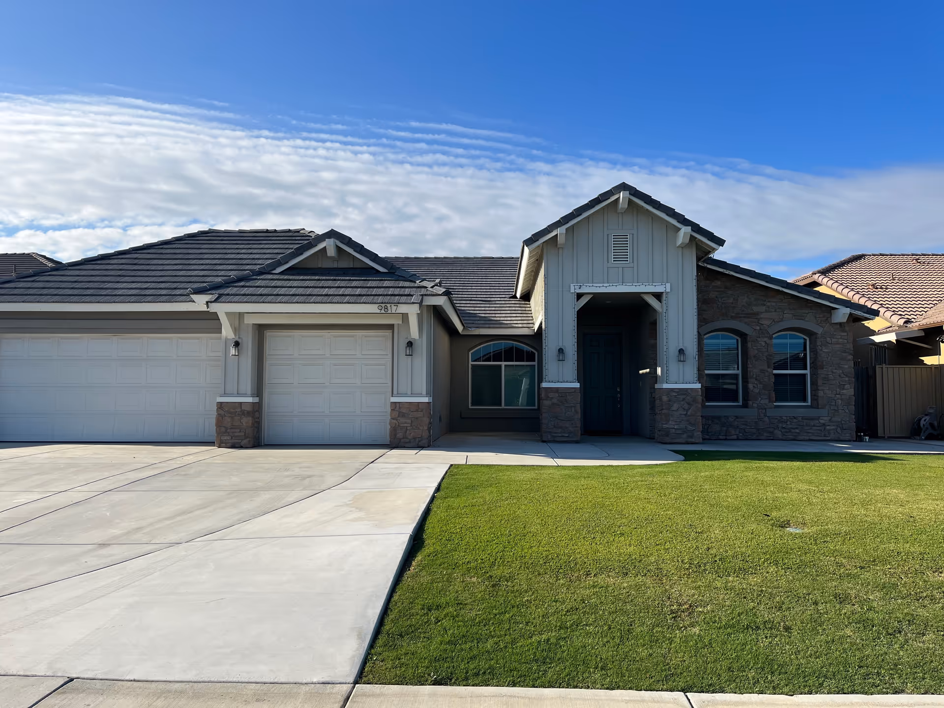 Front exterior view of a single-story residential building with a three-car garage, stone and siding facade, a covered entrance, and a well-maintained green lawn under a blue sky with some clouds.