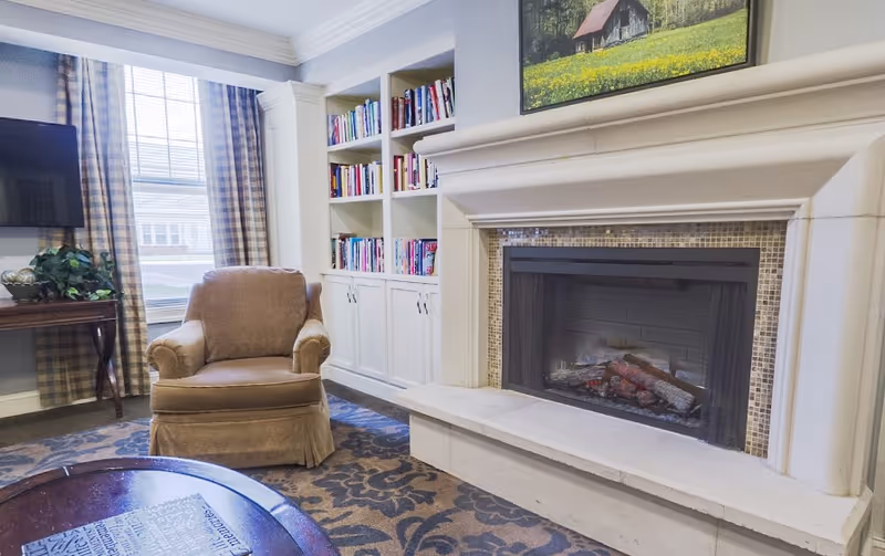 Cozy living room area with a beige armchair, a fireplace with a tiled surround, built-in white bookshelves filled with books, a window with plaid curtains, a wall-mounted TV, and a wooden table with a plant on it.
