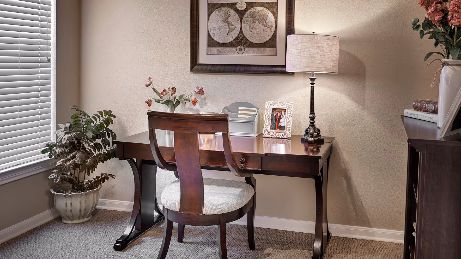 A cozy interior corner featuring a wooden desk with a matching chair. On the desk are a table lamp, a framed photo, a small organizer, and a vase with flowers. A framed world map hangs on the wall above the desk. To the left, there is a window with closed blinds and a potted plant on the floor. To the right, part of a wooden bookshelf with books and a flower arrangement is visible.