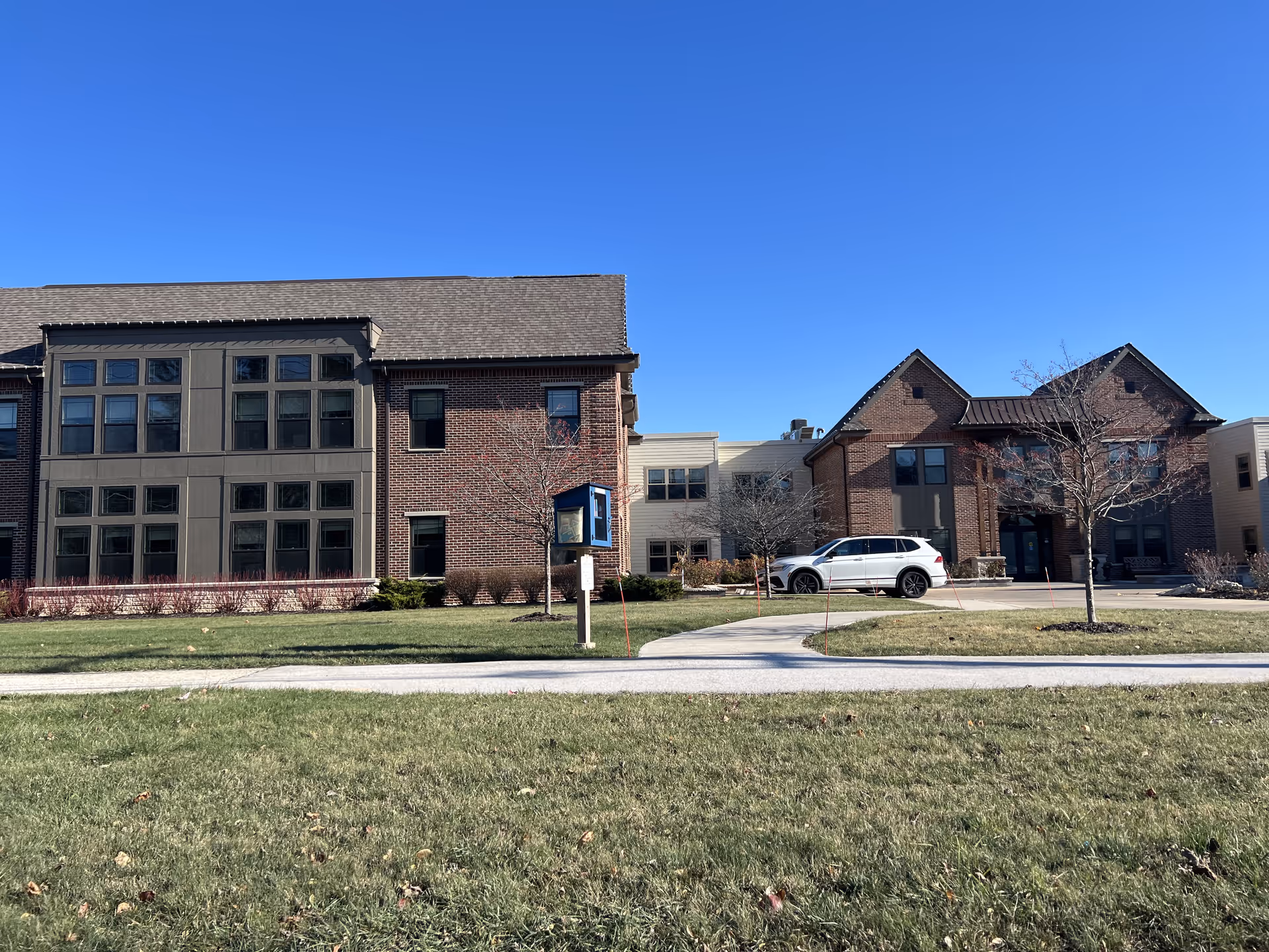 Exterior view of a senior living facility building with brick and beige siding under a clear blue sky. There is a paved walkway, a small lawn with some leafless trees, and a white SUV parked near the entrance.