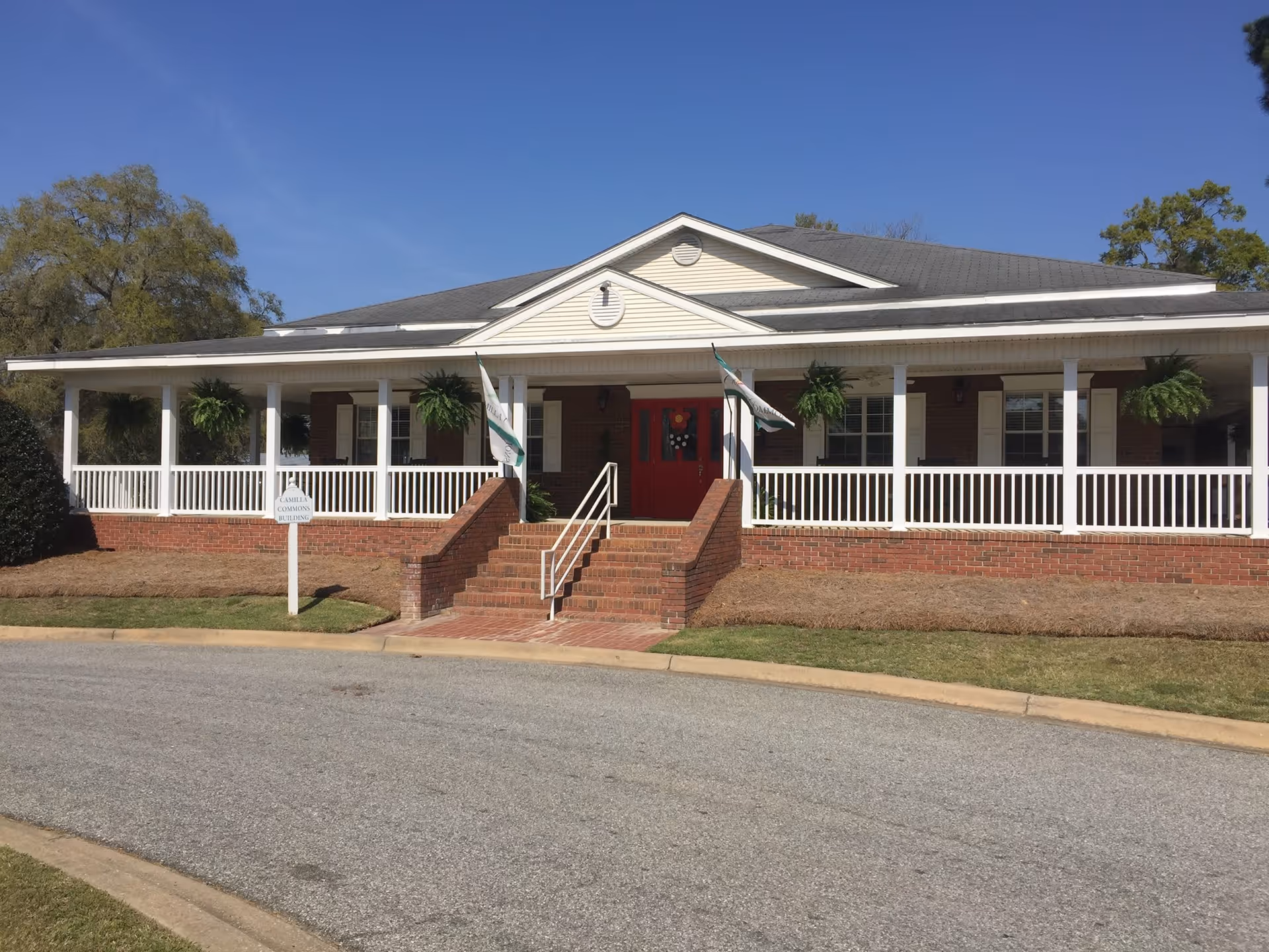 Front exterior of a single-story brick building with a wide white porch, hanging ferns, and red double doors.