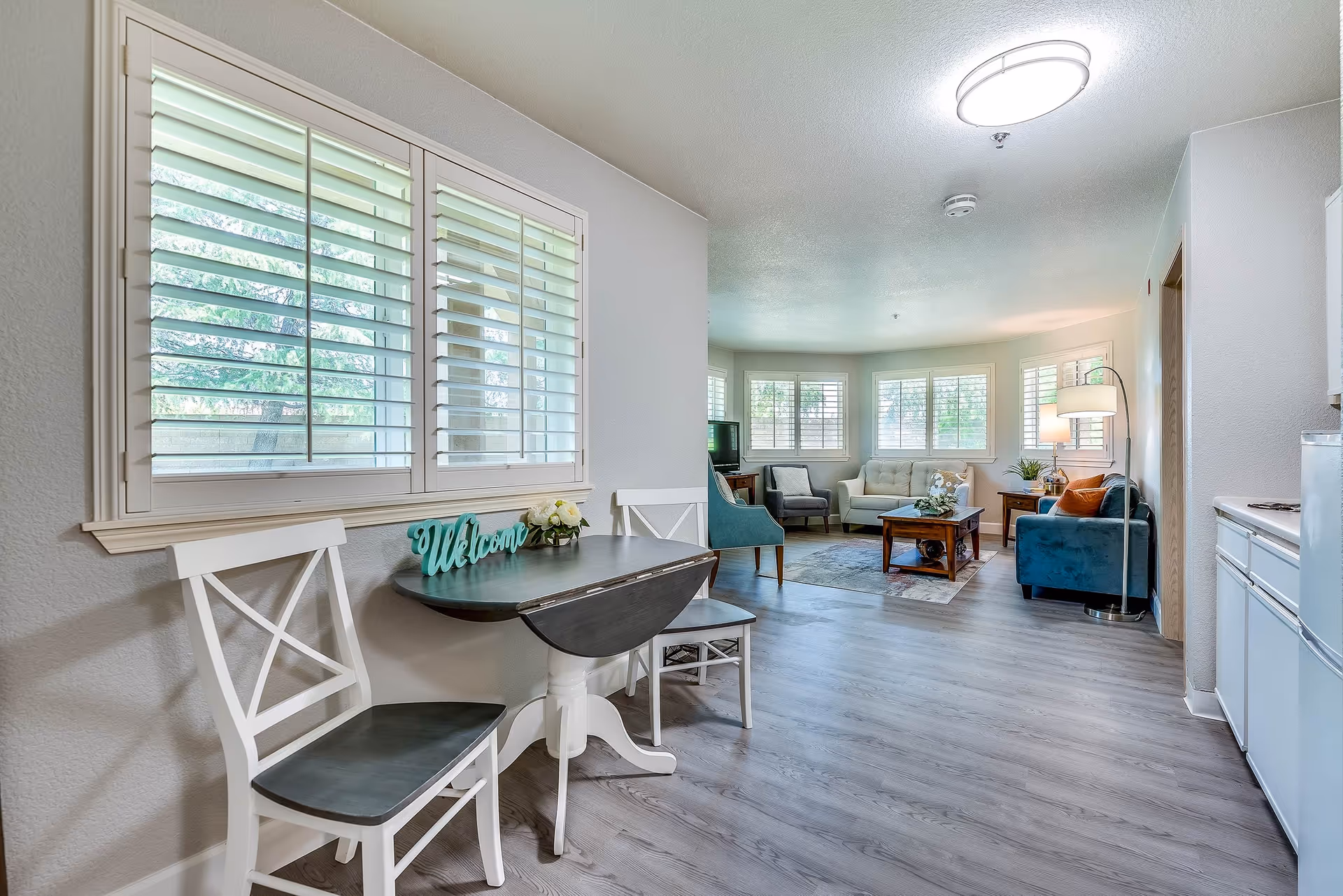 Bright open-plan living and dining area with a small table and chairs by a window and a seating area with sofas and armchairs near large shuttered windows.
