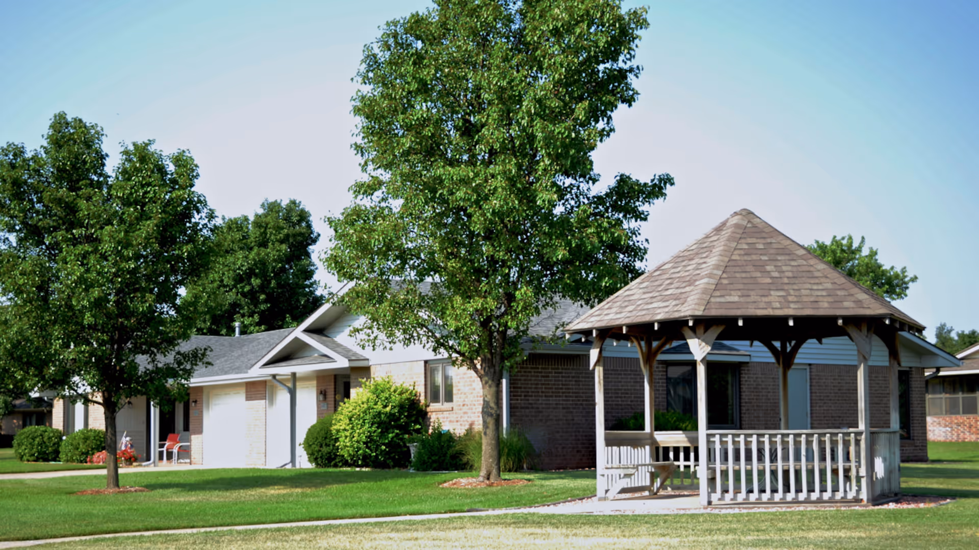 A single-story brick building with a gray roof surrounded by green grass and trees. In the foreground, there is a wooden gazebo with a shingled roof and white railings. The sky is clear and blue.