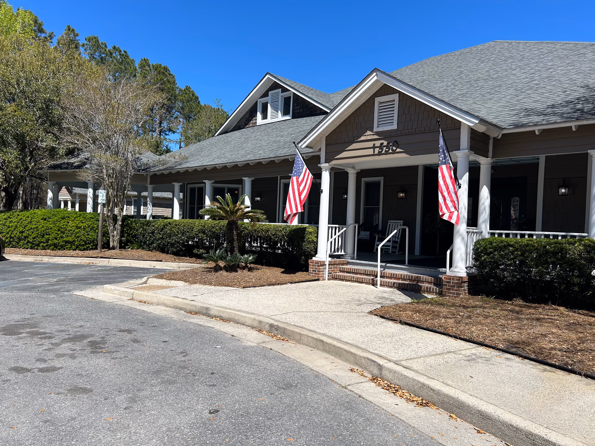 Exterior view of a single-story building with a gray roof and brown siding, featuring a covered porch with white columns and two American flags displayed on either side of the entrance. The building is surrounded by trimmed bushes and trees under a clear blue sky.