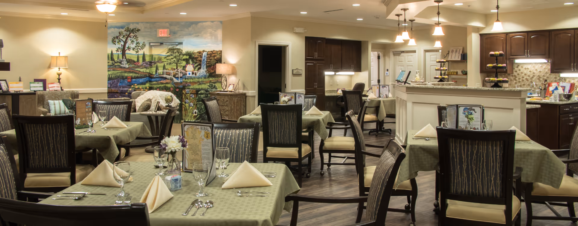 A dining room in a senior living facility with multiple tables covered in green tablecloths, each set with folded napkins, glasses, and silverware. The room features wooden chairs with patterned upholstery, a mural on the far wall depicting a countryside scene, and a kitchen area with cabinets and countertops in the background. Soft lighting from ceiling fixtures and lamps creates a warm atmosphere.
