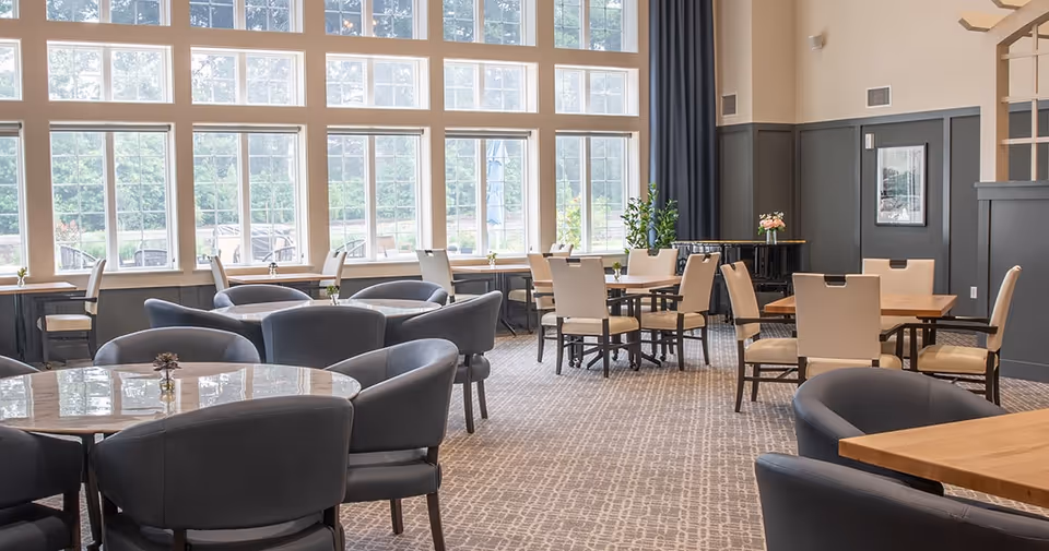 Bright communal dining room with multiple round and rectangular tables, upholstered chairs, and large multi-pane windows.