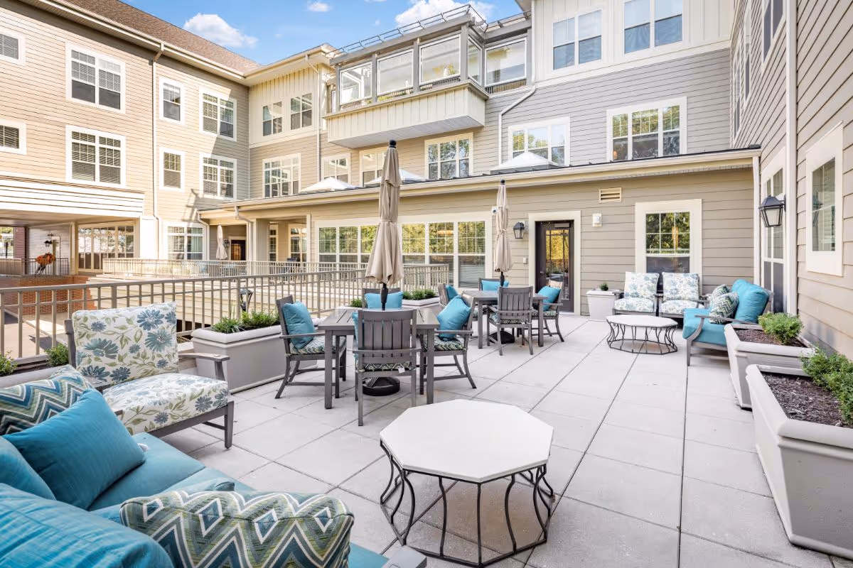 Outdoor patio area at Sunrise of Summit featuring multiple seating arrangements with cushioned chairs and sofas in blue and patterned fabrics, several tables with closed umbrellas, large planters with greenery, and the exterior walls and windows of the building surrounding the patio under a clear blue sky.