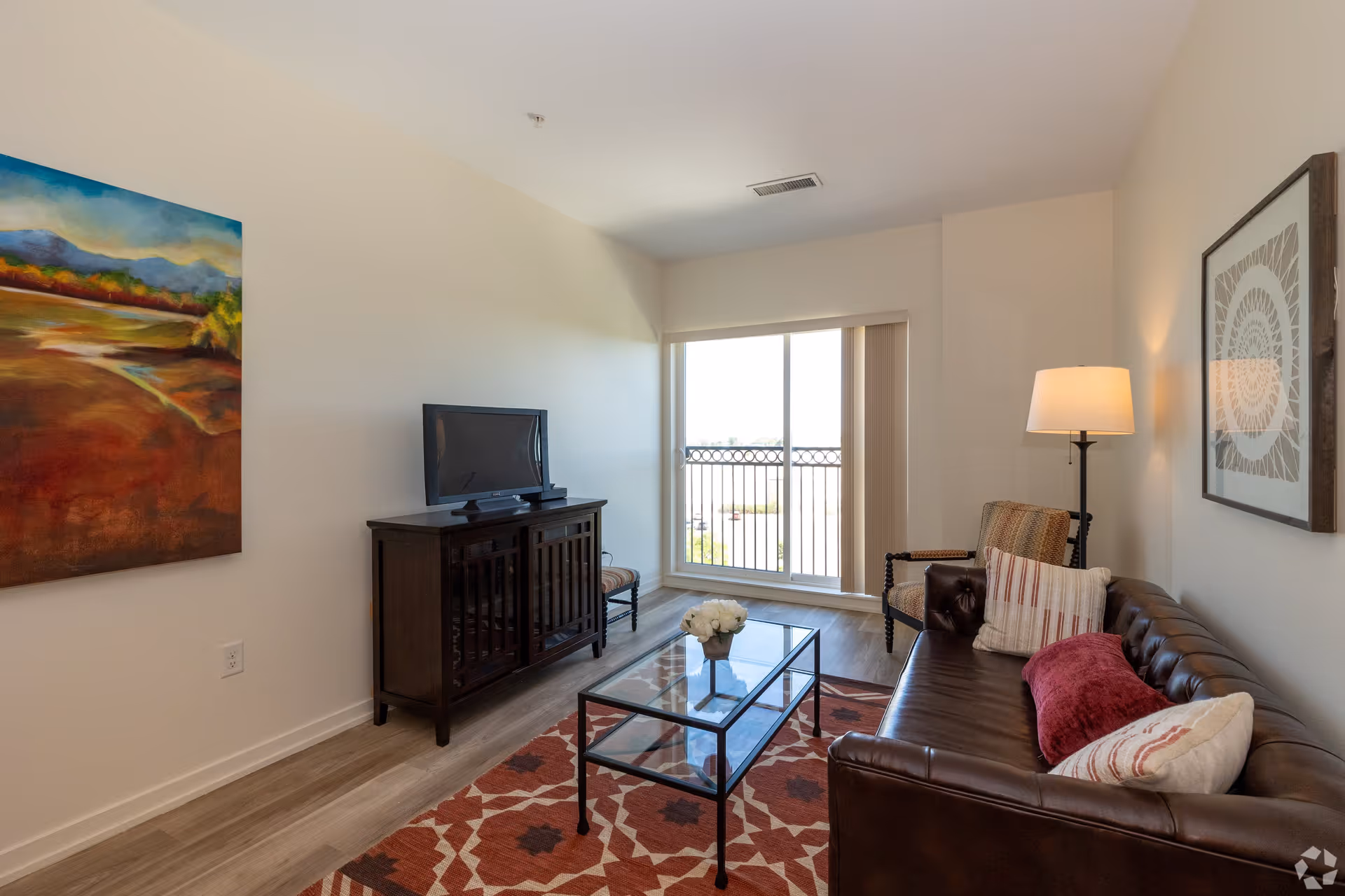 A cozy living room with a brown leather sofa adorned with pillows, a glass coffee table with a flower vase, a patterned rug, a wooden TV stand with a flat-screen TV, a floor lamp, an armchair, and a large window with vertical blinds letting in natural light. There is colorful artwork on one wall and a framed decorative piece on another.