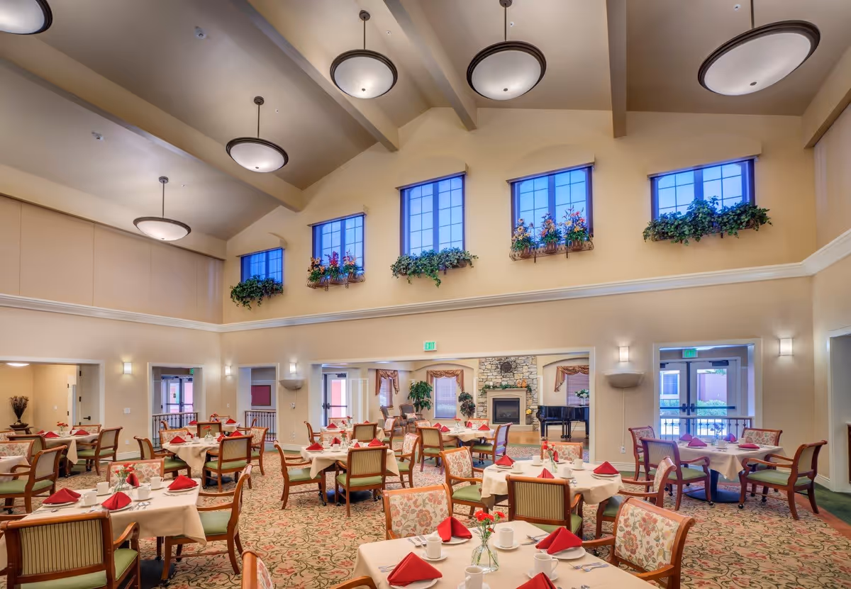 A spacious dining room with multiple tables covered in white tablecloths and set with red folded napkins, white cups, and silverware. The room features high ceilings with large round light fixtures, tall windows adorned with flower boxes and greenery, and a carpeted floor with a floral pattern. In the background, there is a stone fireplace and a black grand piano.