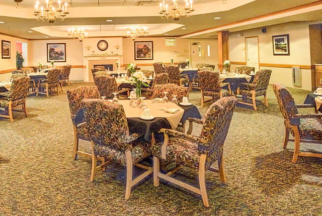 A spacious dining room with multiple tables covered in tablecloths and set with cups and napkins. The room features patterned upholstered chairs, chandeliers hanging from the ceiling, framed artwork on the walls, and a fireplace with a clock above it in the background.