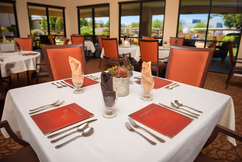 A dining room table set for four with white tablecloth, red menus, silverware, and folded cloth napkins in glasses. The chairs have orange patterned upholstery. Large windows in the background show an outdoor patio area with greenery.