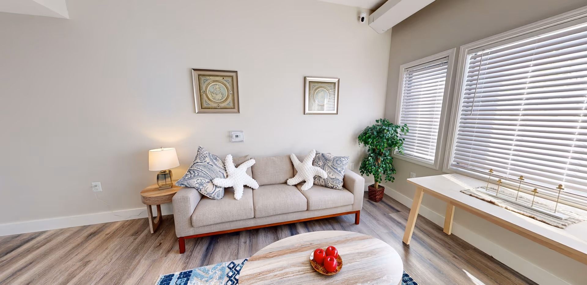 A bright and cozy living room with a beige sofa adorned with patterned pillows and two starfish-shaped cushions. A small wooden side table with a lamp is next to the sofa. Two framed artworks hang on the wall above the sofa. There is a wooden coffee table with a bowl of red apples in front of the sofa. Large windows with white blinds allow natural light to fill the room. A green potted plant is placed near the windows, and a long table with decorative items is positioned against the wall under the windows. The floor has a wood-like finish.
