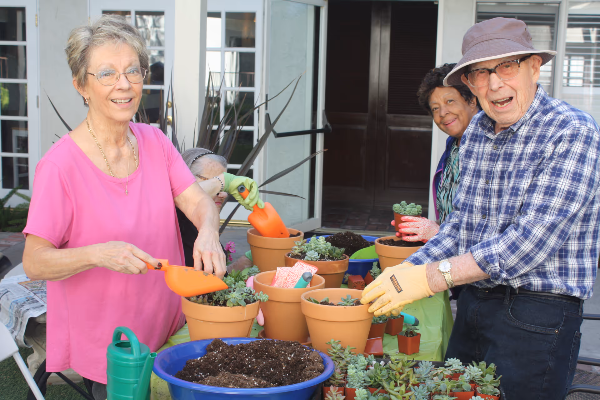 A group of elderly people gardening together outdoors, planting succulents in terracotta pots on a table filled with soil and gardening tools. They are smiling and appear to be enjoying the activity.