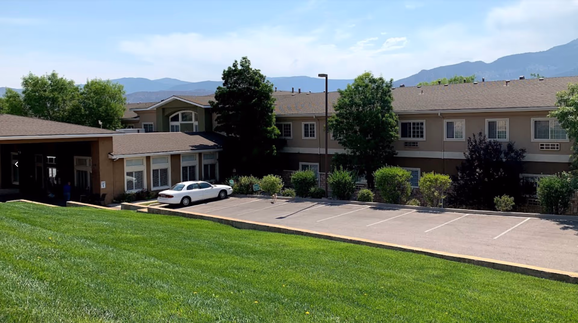 Exterior view of a two-story assisted living facility building with a parking lot in front, surrounded by green grass, trees, and bushes, with mountains visible in the background under a partly cloudy sky.