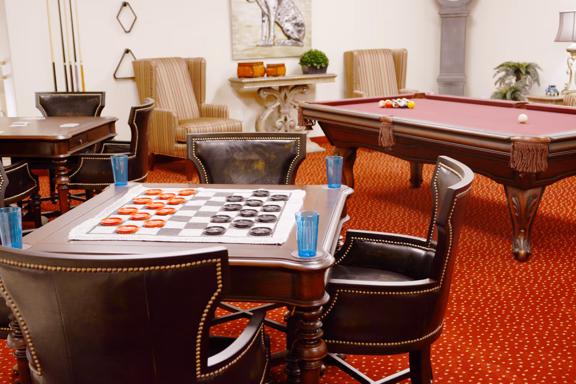 A cozy game room with a checkers board set up on a wooden table surrounded by four leather chairs. In the background, there is a pool table with balls arranged for a game, two striped armchairs, a decorative console table with plants and pottery, and a red patterned carpet covering the floor.
