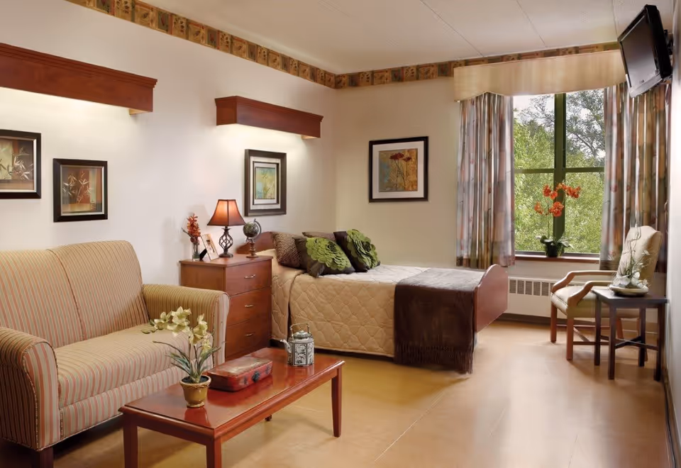 A cozy senior living bedroom with a single bed covered in a beige quilt and green decorative pillows. Next to the bed is a wooden nightstand with a lamp, a small globe, and framed photos. A striped loveseat sits beside a wooden coffee table with a potted plant and decorative box. The room has framed artwork on the walls, a large window with floral curtains letting in natural light, and a wall-mounted TV in the corner. A wooden chair and small side table with an orchid plant are near the window.