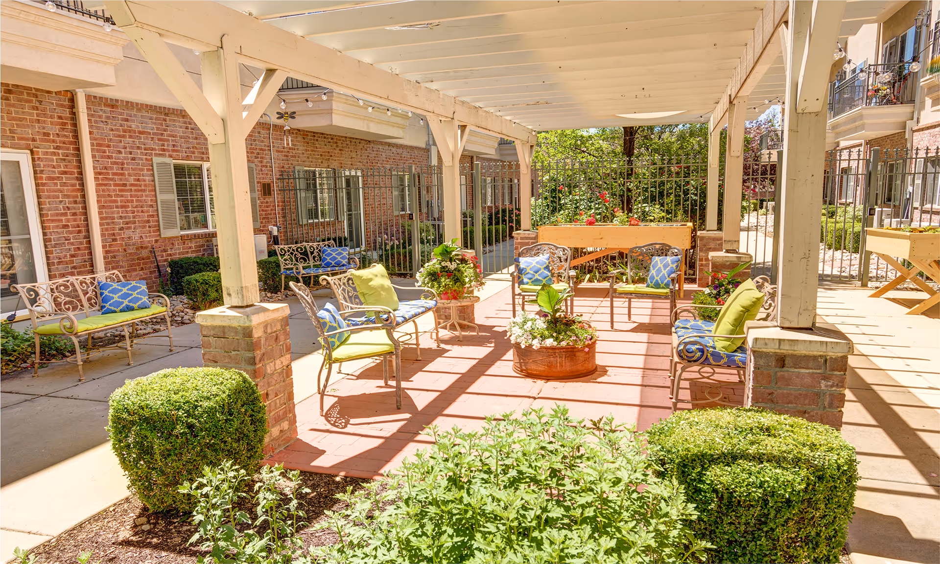 Shaded outdoor courtyard patio with a pergola, metal chairs with colorful cushions, potted plants, and trimmed shrubs.