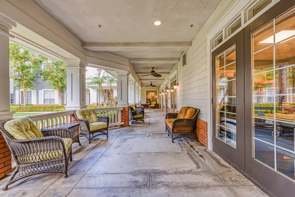 Covered outdoor patio area with wicker chairs featuring green and orange cushions, ceiling fans, and wall-mounted lantern lights. The patio overlooks a landscaped garden with trees and shrubs, and there are glass doors leading inside the building.