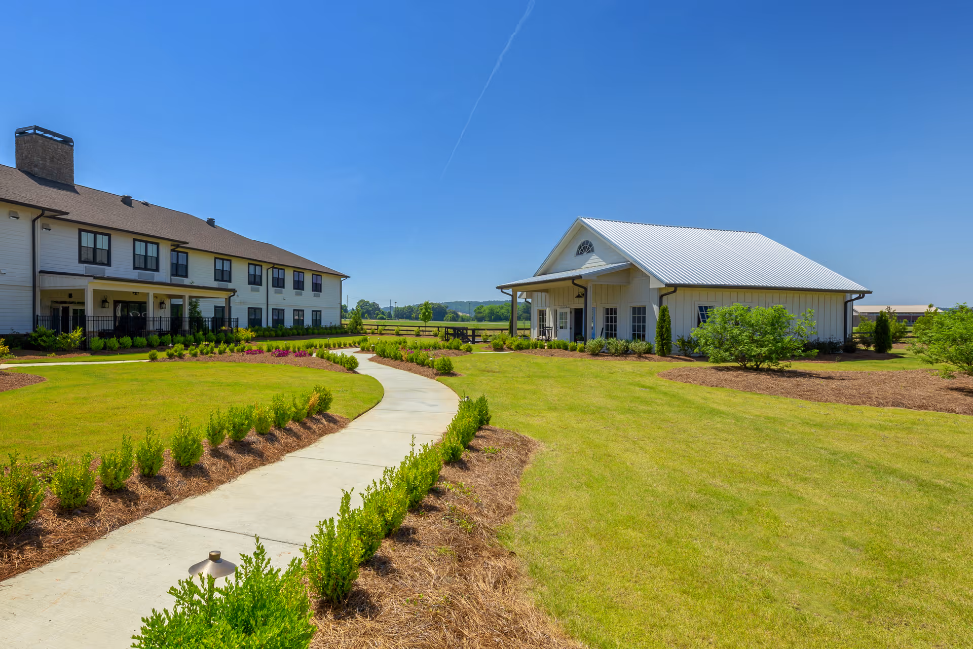 A curved concrete path through manicured lawns leading to two light-colored residential buildings under a clear blue sky.