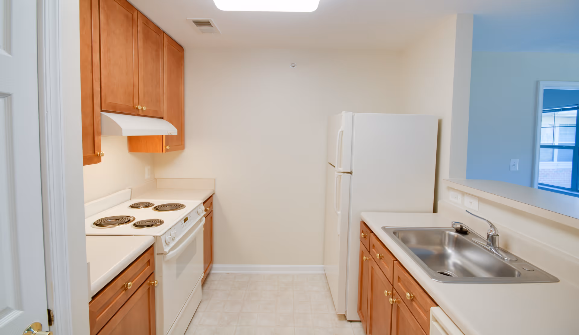A small kitchen with wooden cabinets, a white electric stove with four coil burners, a white refrigerator, a stainless steel sink, and beige countertops. The floor is covered with light-colored tiles and the walls are painted off-white. There is a pass-through window opening to another room.
