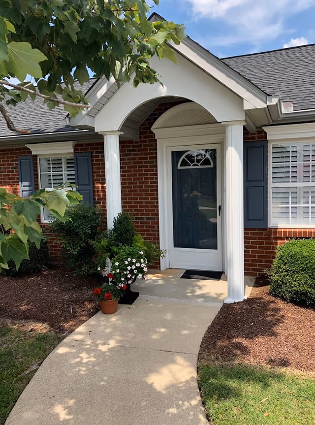Front entrance of a brick retirement home with a covered porch supported by white columns, a dark entry door, shuttered windows and potted flowers by the walkway.