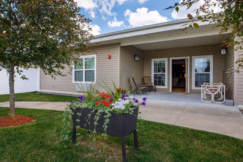 Outdoor patio area of a senior living facility with beige siding, two windows, and an open door leading inside. The patio has chairs and a small table, with a flower planter filled with colorful flowers in the foreground and a tree to the left. The sky is partly cloudy.