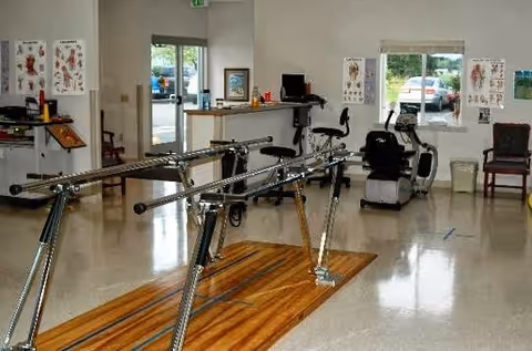 Physical therapy room with parallel bars for walking exercises, a recumbent exercise bike, chairs, anatomical charts on the walls, and large windows showing a parking lot outside.