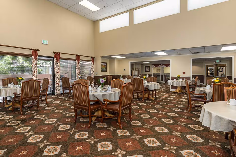 Spacious dining room with round tables set with white tablecloths, wooden chairs, and floral centerpieces under high ceilings.