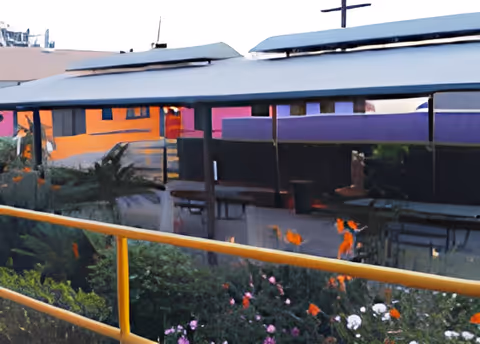 Outdoor area with a yellow railing in the foreground, flowers and greenery below it, and a covered walkway or shelter with a reflective surface showing distorted reflections of nearby buildings and plants.