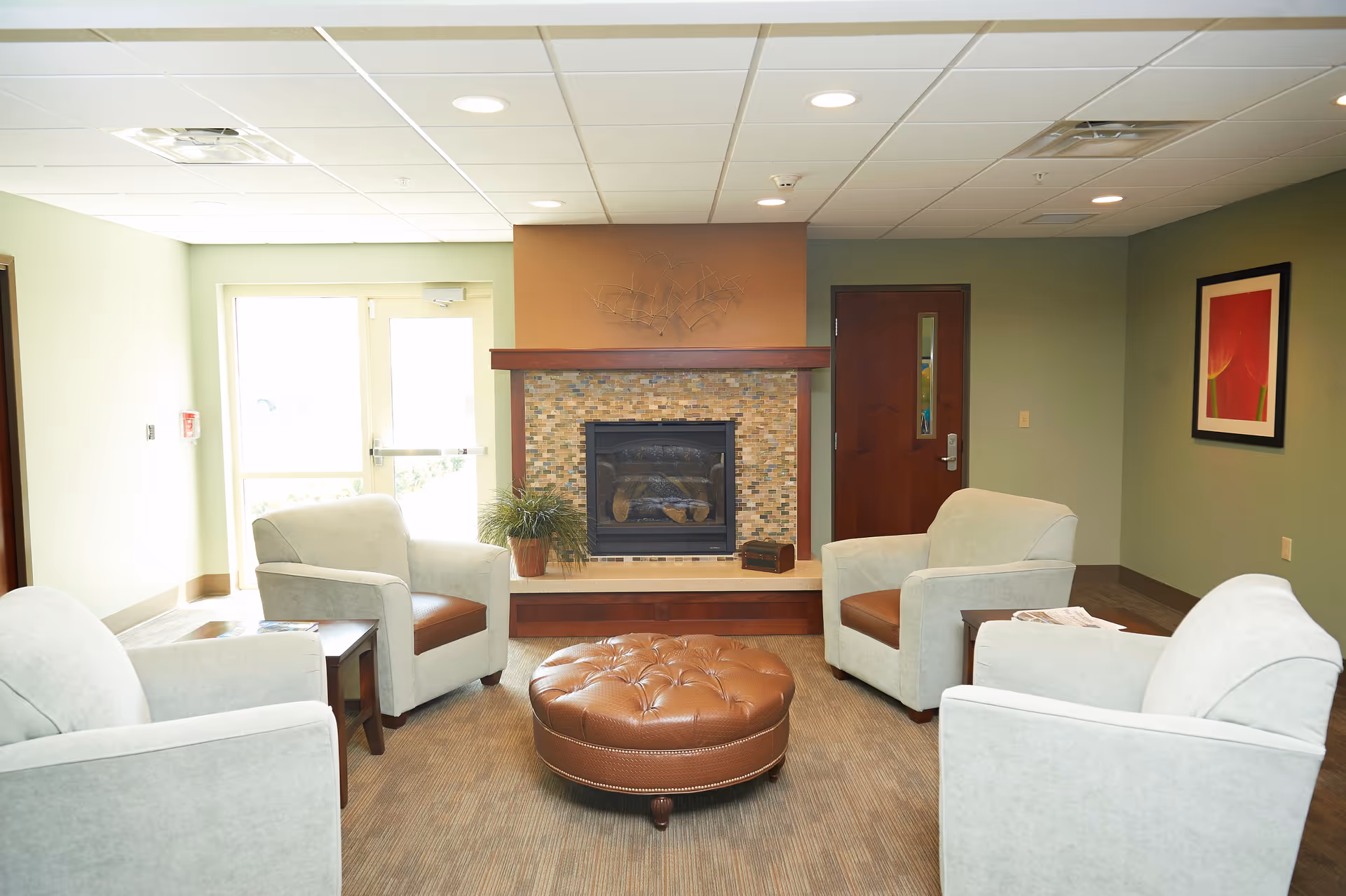 A cozy senior living lounge with four armchairs arranged around a tufted ottoman facing a fireplace.