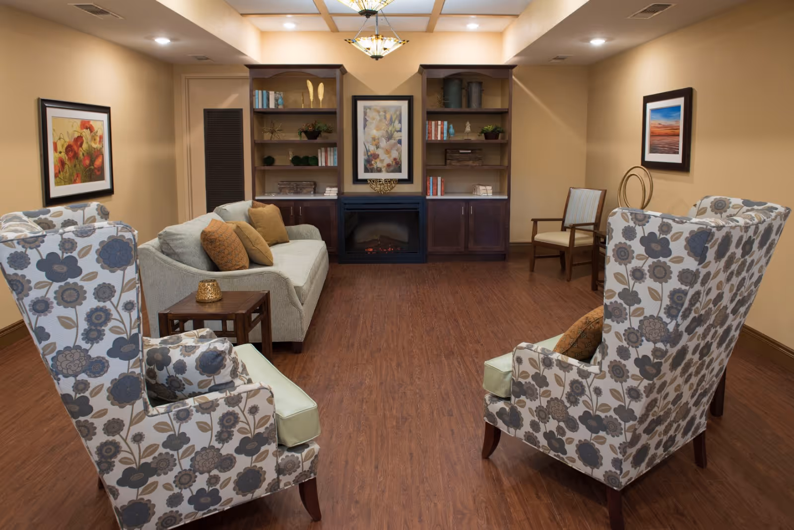 A cozy living room with two floral-patterned armchairs facing a light gray sofa adorned with brown and orange cushions. There is a small wooden side table with a candle holder between the sofa and one armchair. The room features wooden flooring, beige walls, and built-in dark wood shelves flanking a fireplace with a framed floral painting above it. Additional framed artwork hangs on the walls, and a wooden chair with light upholstery is positioned near the right wall.