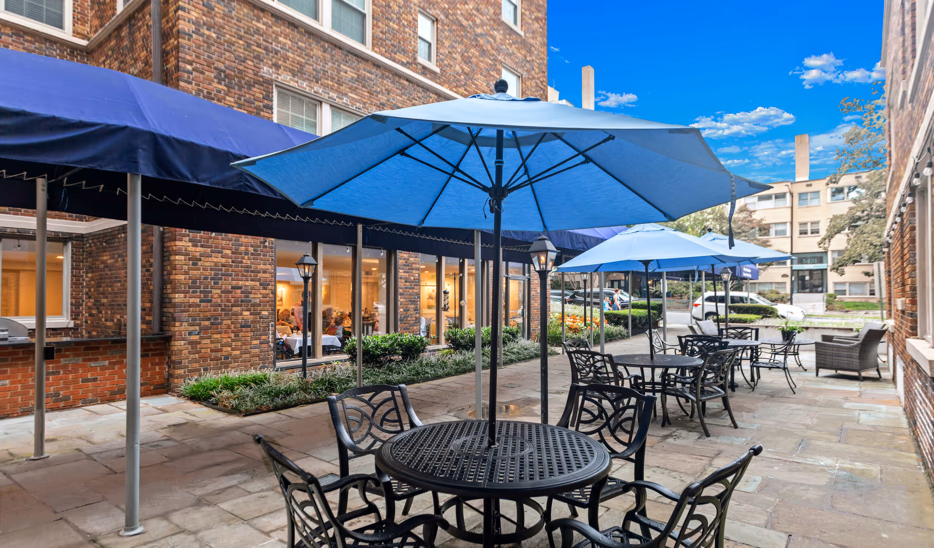 Outdoor courtyard patio with metal tables, chairs, and blue umbrellas beside a brick building.