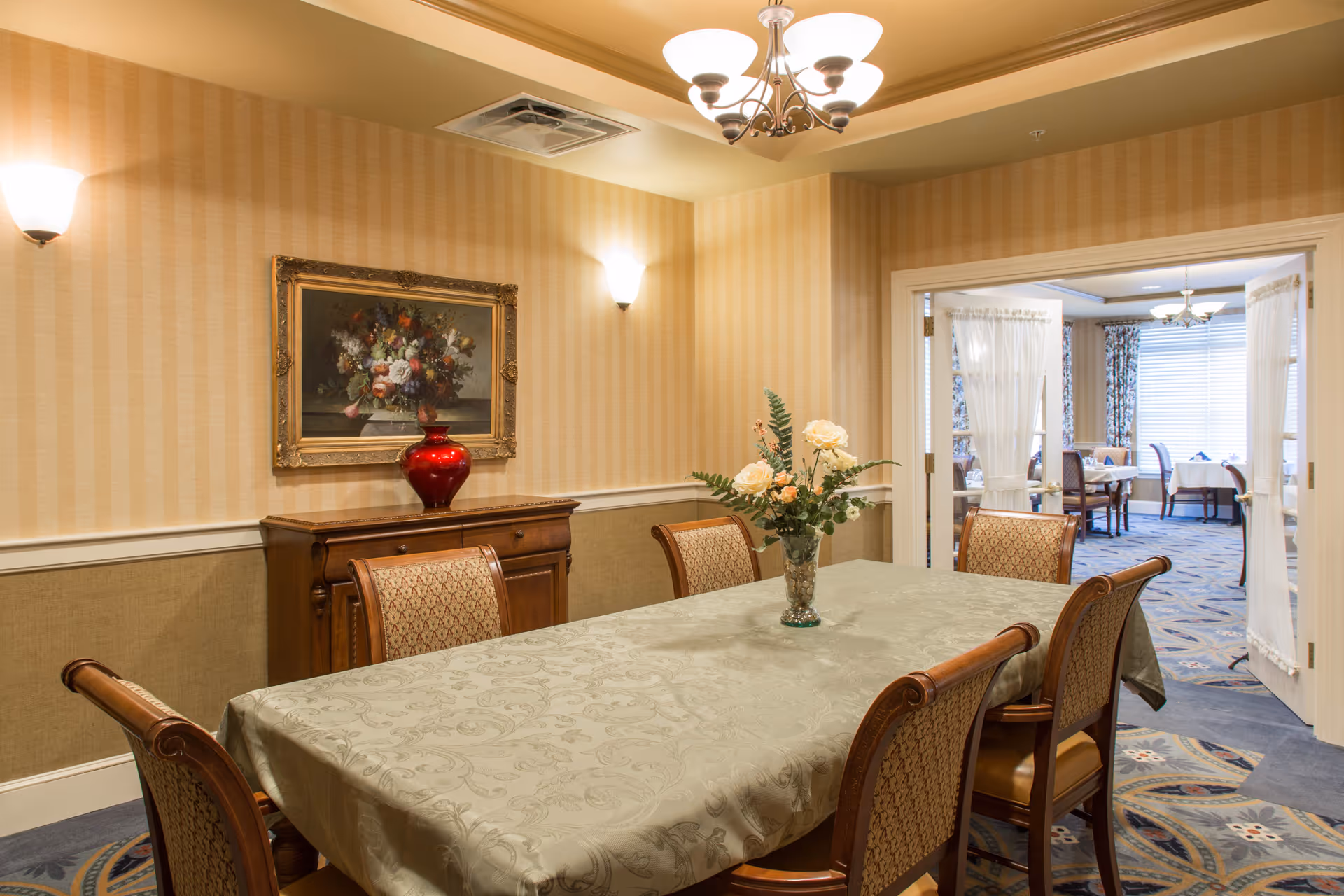A dining room with a rectangular table covered with a patterned tablecloth and six upholstered wooden chairs. A vase with flowers is placed in the center of the table. The walls have striped wallpaper with two wall sconces and a framed floral painting above a wooden sideboard with a red vase. Double doors open to another dining area with tables and chairs near large windows with blinds and floral curtains.