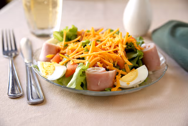 A glass plate with a fresh salad consisting of lettuce, shredded cheddar cheese, tomato pieces, rolled slices of ham, and quartered hard-boiled eggs. The plate is on a beige tablecloth with a fork and knife to the left and a glass of a light-colored beverage in the background.