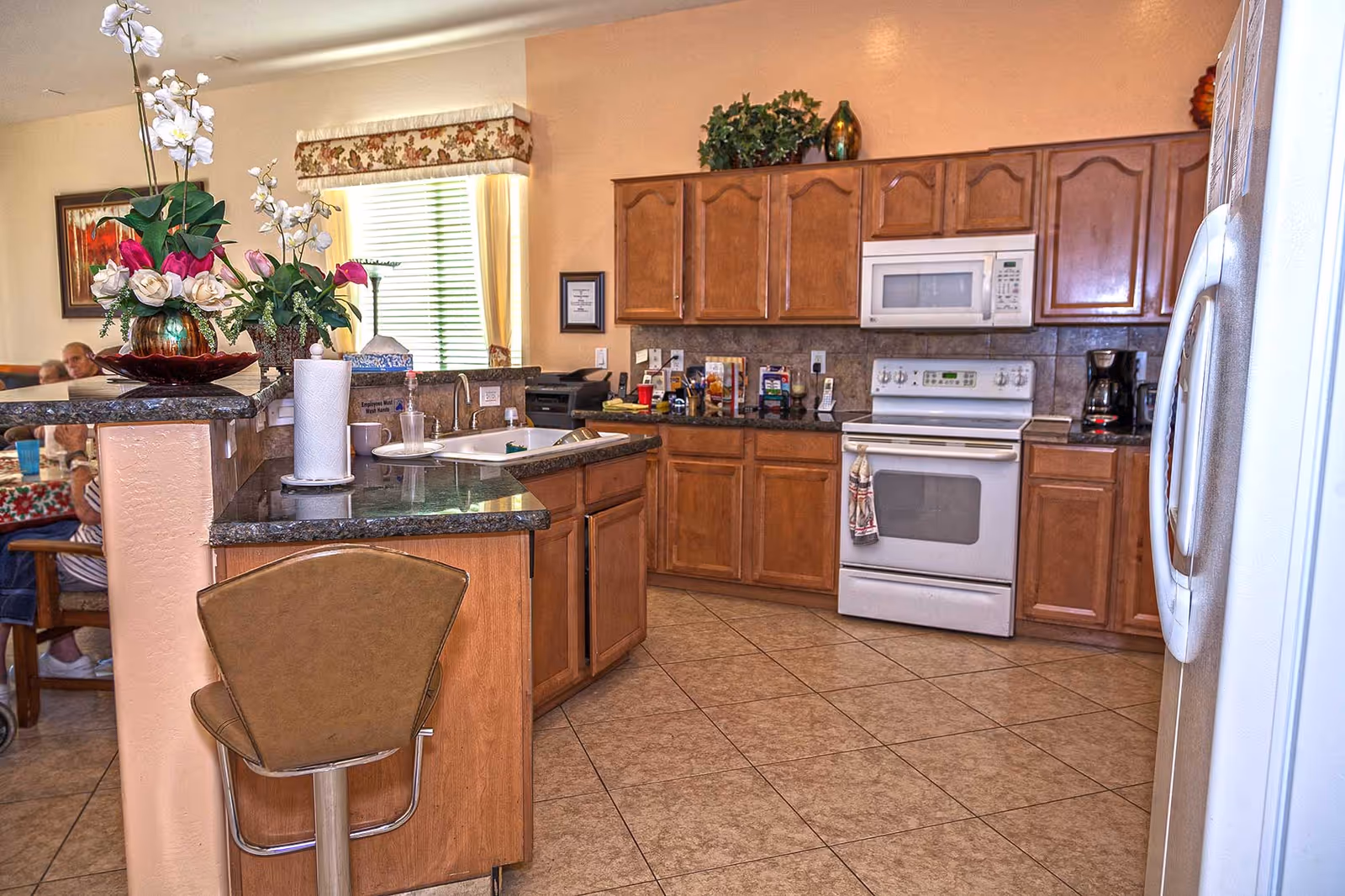 Bright kitchen with wooden cabinets, a white stove and microwave, a center island with sink and bar stool, floral arrangement on the counter, and a dining area with seated residents visible in the background.