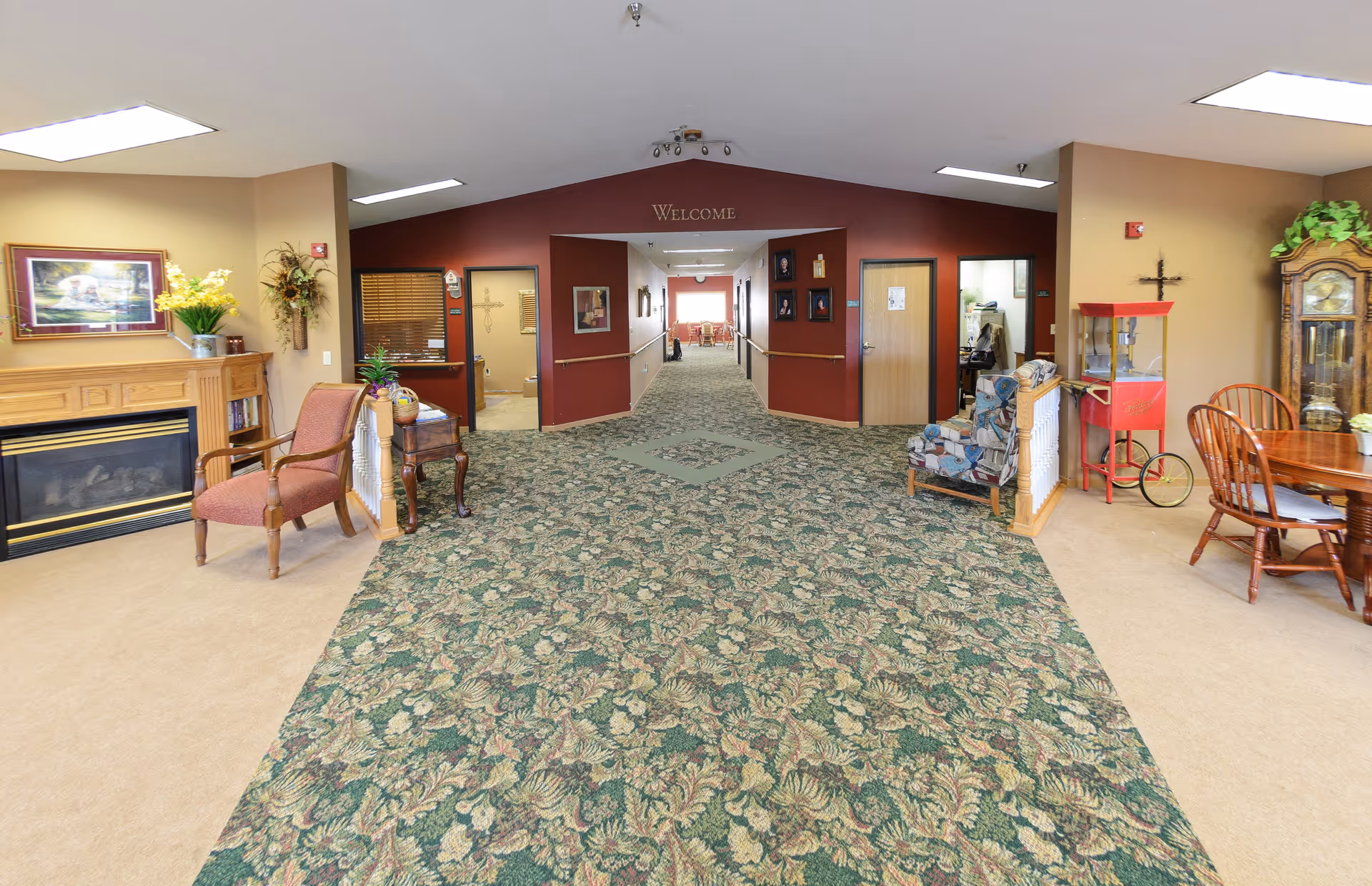 Carpeted senior living facility common area with chairs, a fireplace, a small dining table, and a central hallway under a 'Welcome' arch.