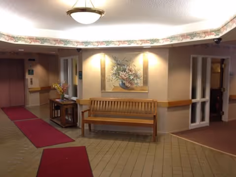 Interior hallway of a senior living facility with beige walls, tiled floor, and red carpet runners. A wooden bench is placed against the wall beneath a framed floral painting. To the left, there is an elevator with closed doors, and to the right, doorways lead to other rooms. A small table with a flower arrangement is next to the bench.