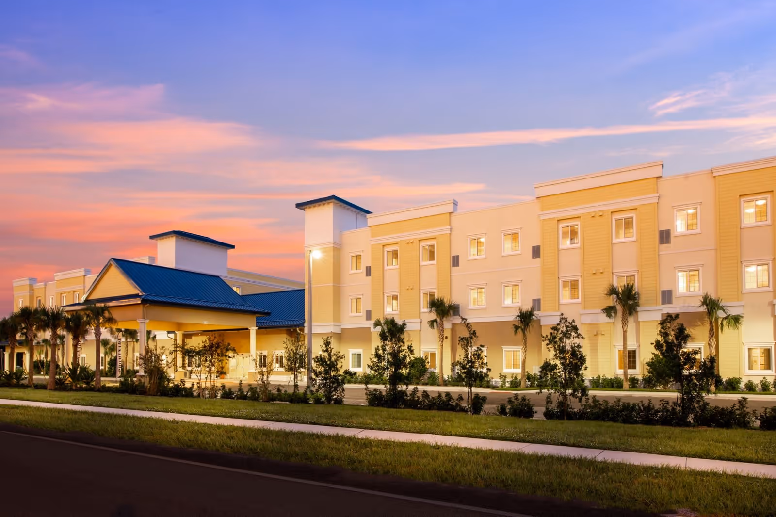 Exterior view of The Promenade senior living facility at sunset, showing a large, multi-story building with a covered entrance, palm trees, and landscaped greenery along the sidewalk.