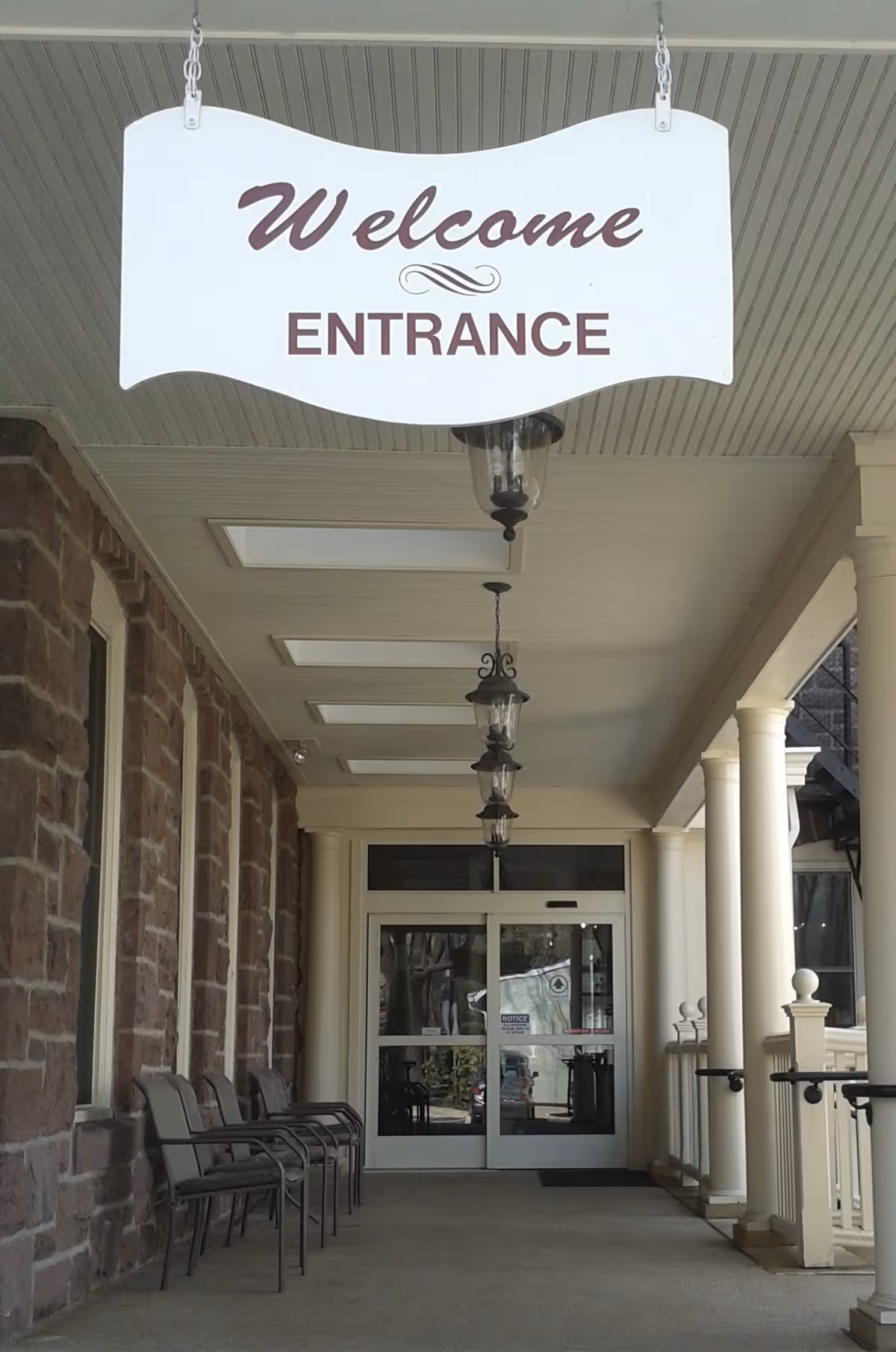 Covered entrance area with a hanging sign that reads 'Welcome ENTRANCE'. There are several chairs lined up against a stone wall on the left side and white columns on the right side. Glass double doors are at the end of the walkway.