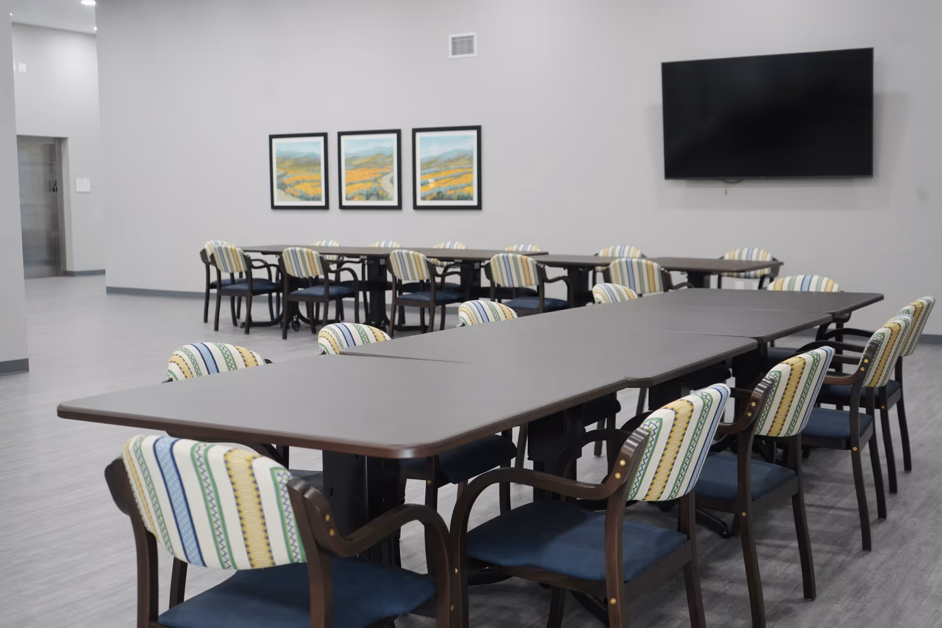 A large room with two long tables arranged in a row, surrounded by chairs with patterned backs and blue seats. On the wall, there are three framed landscape paintings and a large flat-screen TV mounted. The floor is light gray, and the walls are painted a light neutral color. An elevator door is visible in the background.